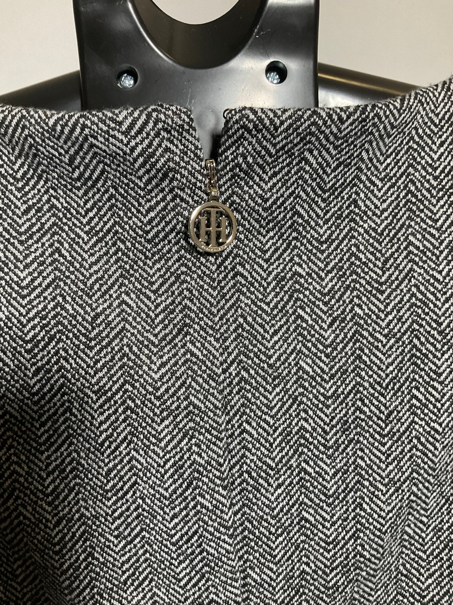 Close-up of a gray and white herringbone tweed fabric with a small circular metal logo charm featuring the letters 'HH'.