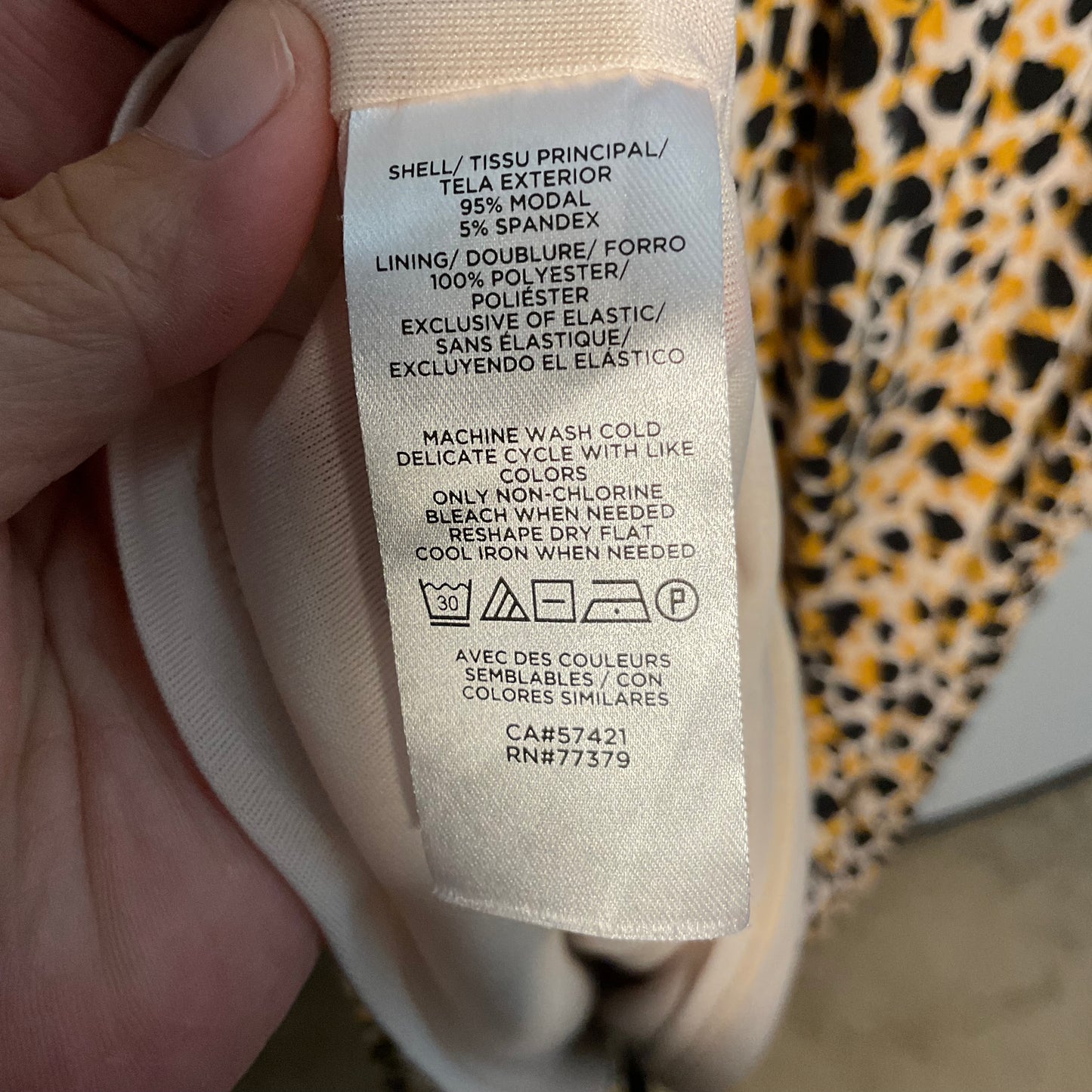 A close-up of a sewn-on fabric care label inside a garment, held by a hand. The label contains product details in English and French, including material composition and care instructions, with a small care symbol block at the bottom.