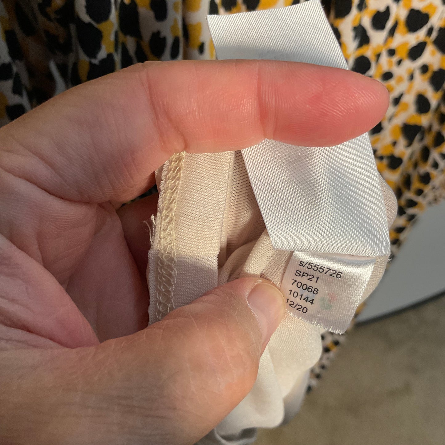 A close-up of a hand holding a garment's interior label, showing size information. The fabric is a light cream color, and the background shows a yellow, black, and white leopard print garment.