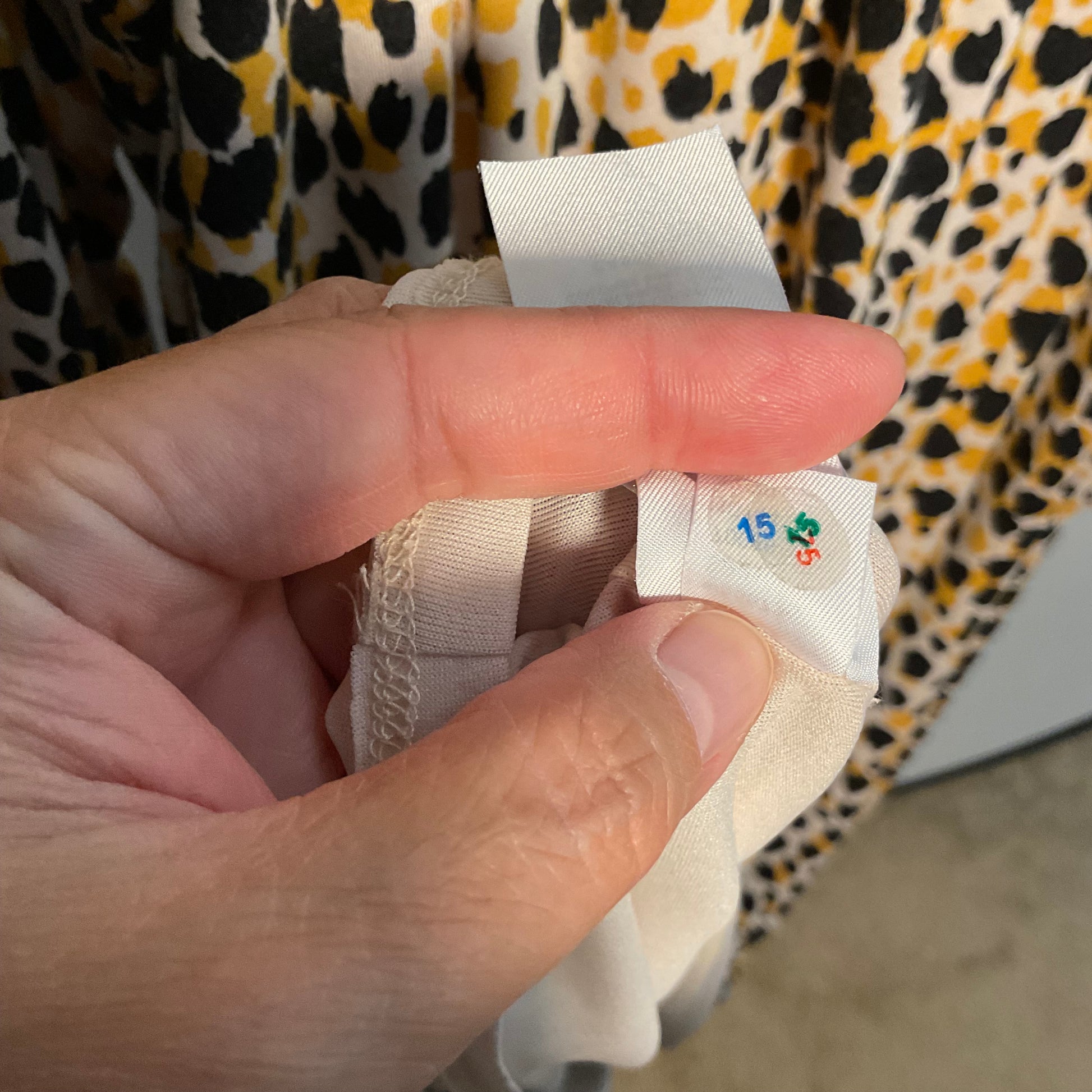 A close-up of a hand holding a beige fabric garment tag with the size '15' printed on it, set against a yellow, black, and white leopard print fabric background.