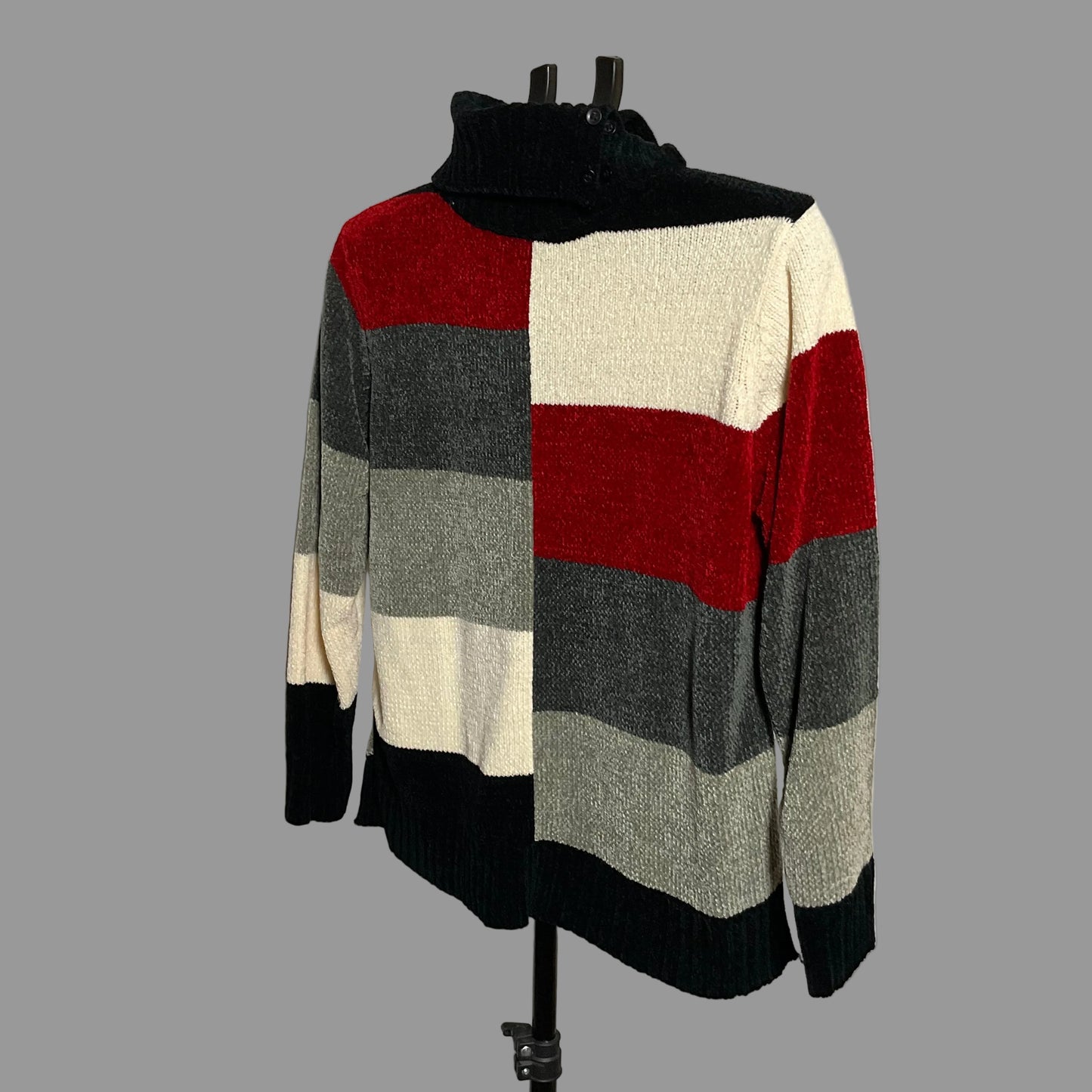 A color-block knit sweater displayed on a black hanger stand against a plain light gray wall. The sweater features a high black ribbed mock neck, horizontal stripes in red, cream, dark gray, and light gray, with black ribbed cuffs and hem.