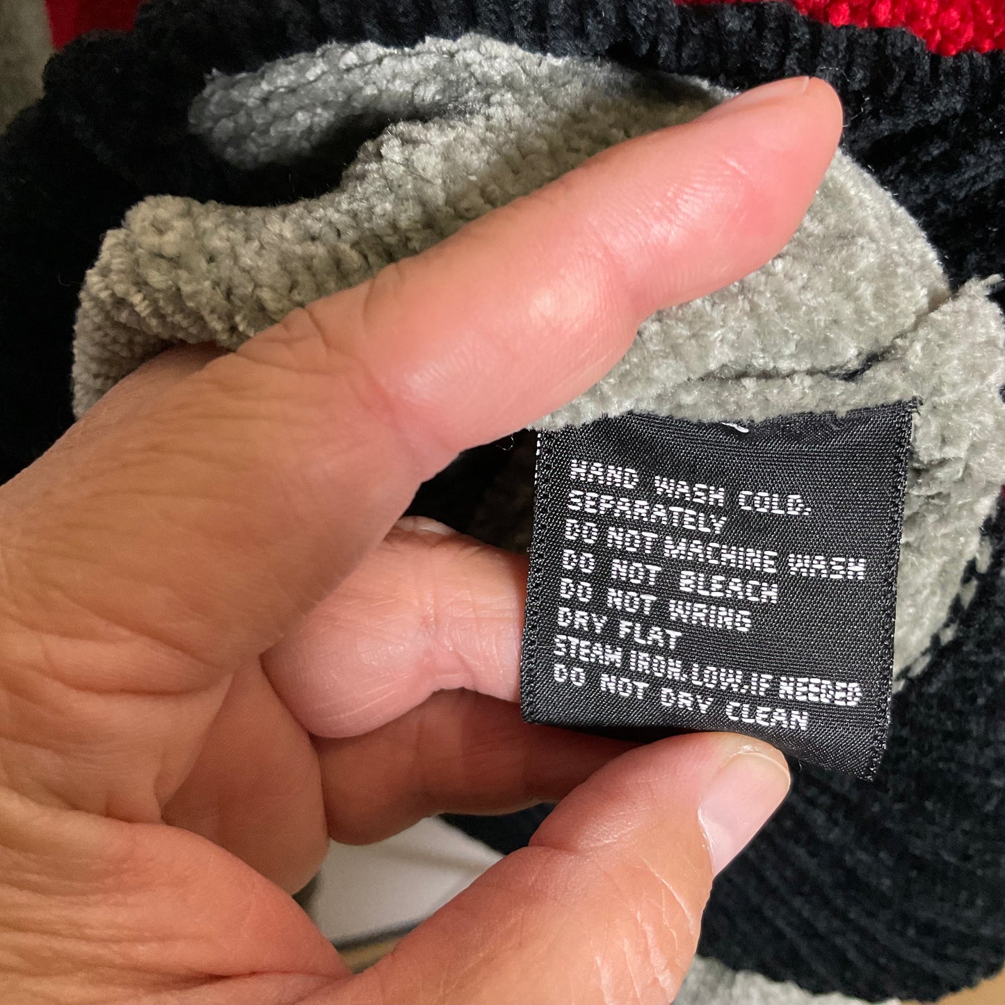 A close-up of a hand holding a black knit beanie, showing a sewn-in care label with white text that reads 'HAND WASH COLD. SEPARATELY.