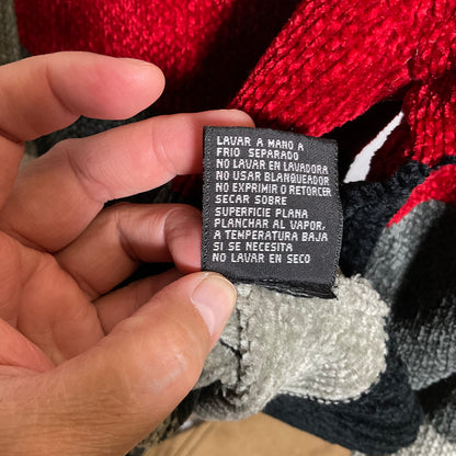A close-up of a hand holding a black fabric care label with white Spanish text, set against a red and black textured knit garment.