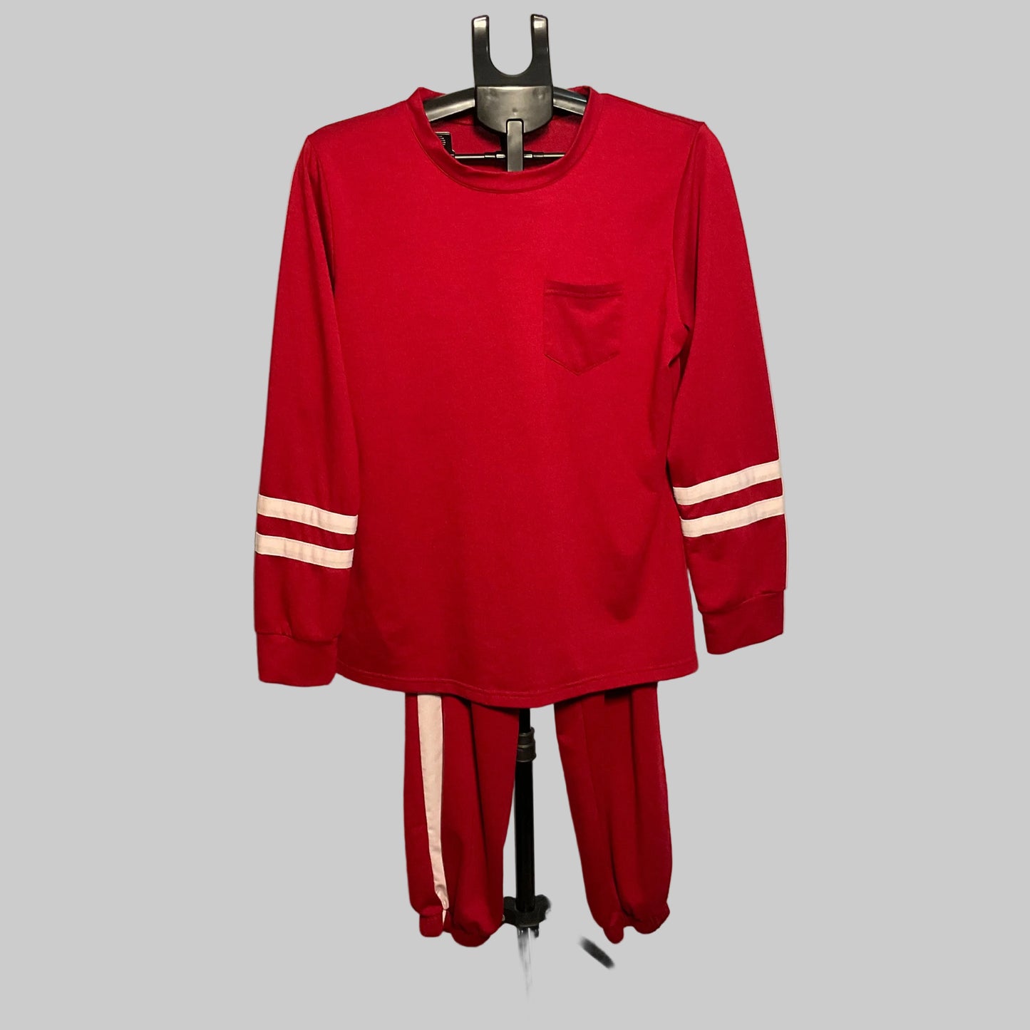 A red two-piece tracksuit set consisting of a long-sleeve shirt and matching pants, displayed on a hanger stand in front of a circular studio backdrop. The shirt features a chest pocket and white stripes on the sleeves, while the matching pants have white stripes down the side.