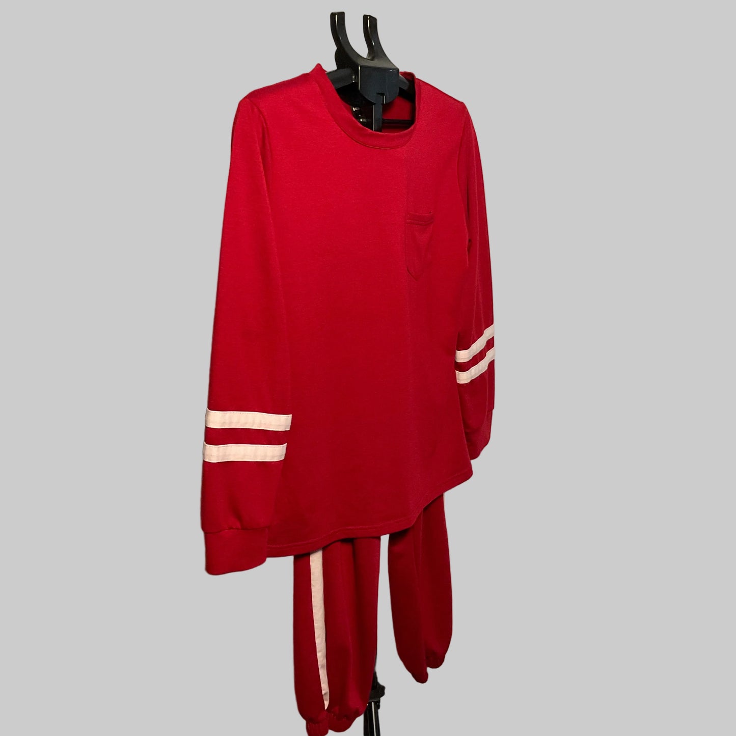 A red two-piece tracksuit set consisting of a long-sleeve top and matching pants, displayed on a black hanger stand. The top features a small chest pocket and white stripes on both sleeves, while the matching pants have a white stripe down the side.