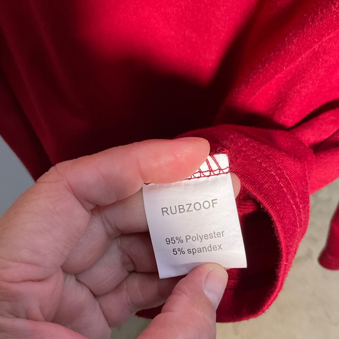 A close-up of a hand holding a white fabric tag that reads 'RUBZOOF 95% Polyester 5% spandex' against a bright red garment.