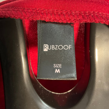 Close-up of a black fabric brand tag reading RUBZOOF and SIZE M, attached inside the collar of a red garment, with a dark plastic hanger visible.