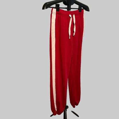 Red sweatpants with a white side stripe and white drawstring, hanging on a black hanger against a white circular backdrop.