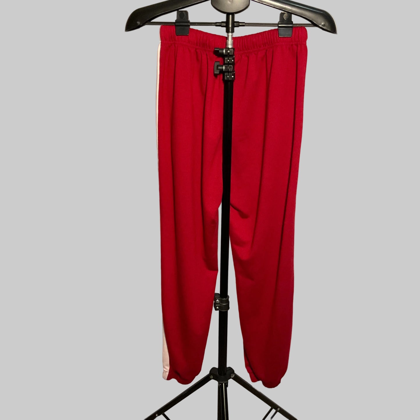 Red athletic sweatpants with a white side stripe, hanging on a black hanger stand in front of a white circular studio backdrop.