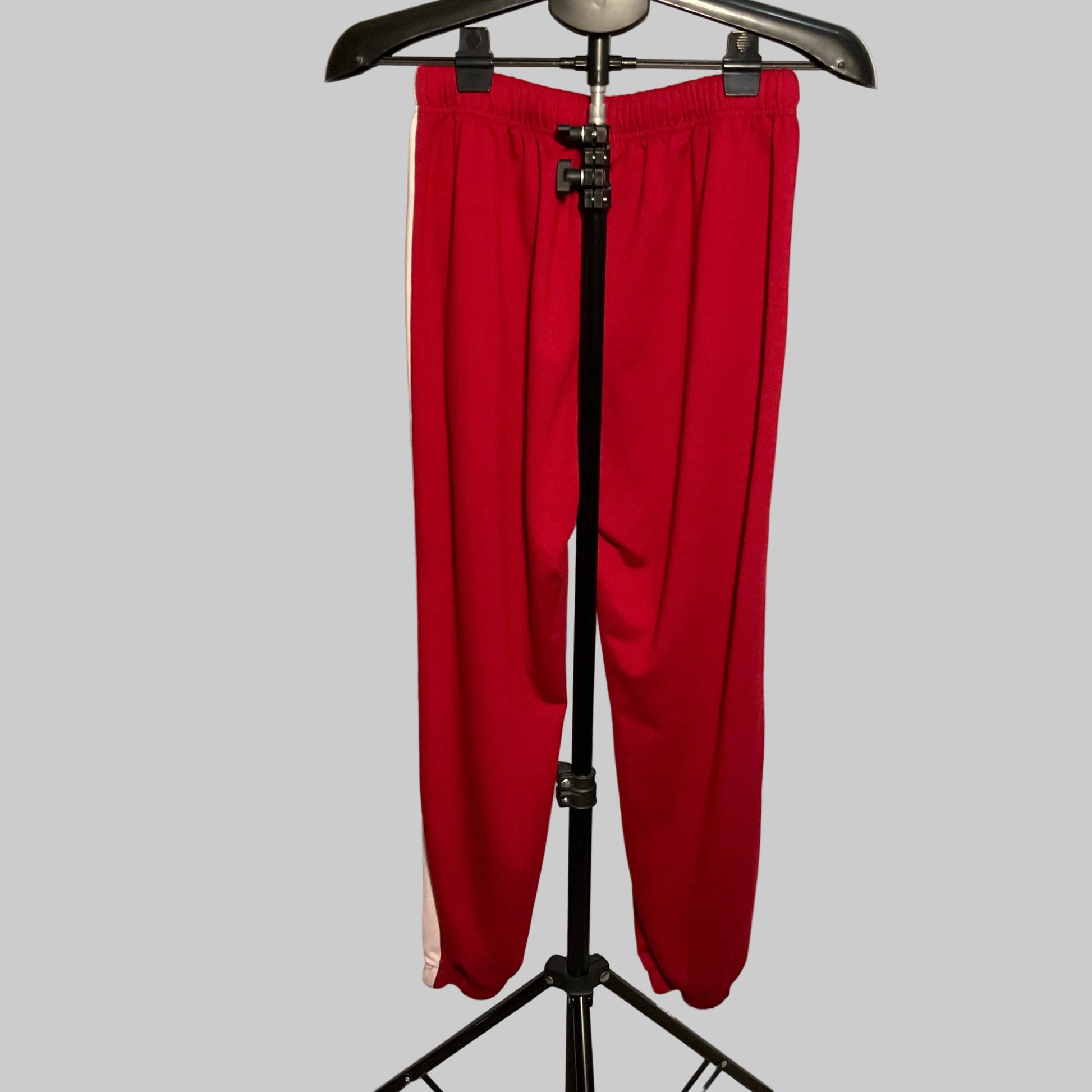 Red athletic sweatpants with a white side stripe, hanging on a black hanger stand in front of a white circular studio backdrop.