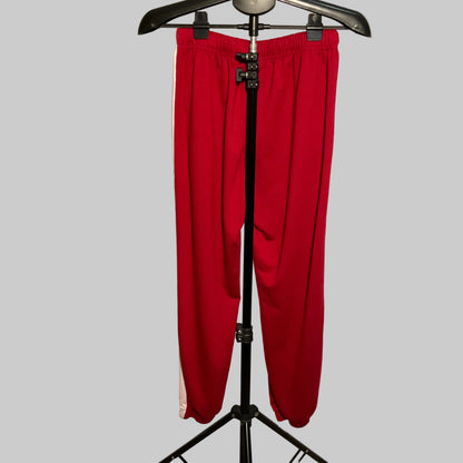 Red athletic sweatpants with a white side stripe, hanging on a black hanger stand in front of a white circular studio backdrop.