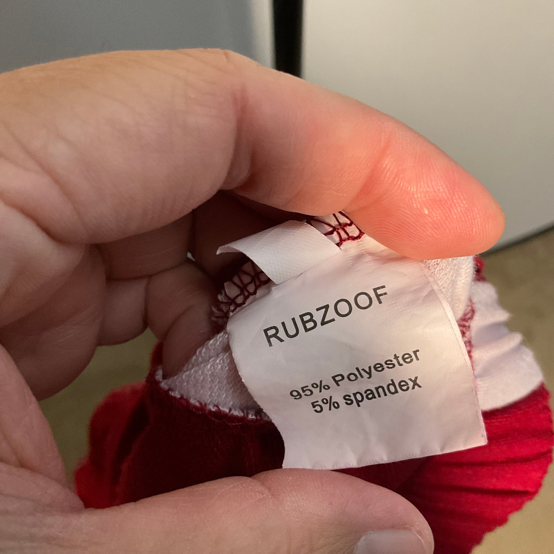 A close-up of a hand holding the inner neck label of a red knit pet sweater. The white fabric label is printed with the brand name 'RUBZOOF' and material composition '95% Polyester 5% spandex'.