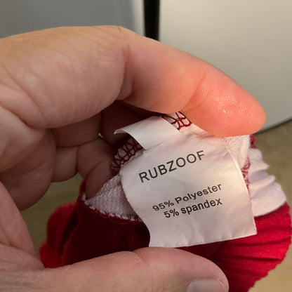 A close-up of a hand holding the inner neck label of a red knit pet sweater. The white fabric label is printed with the brand name 'RUBZOOF' and material composition '95% Polyester 5% spandex'.