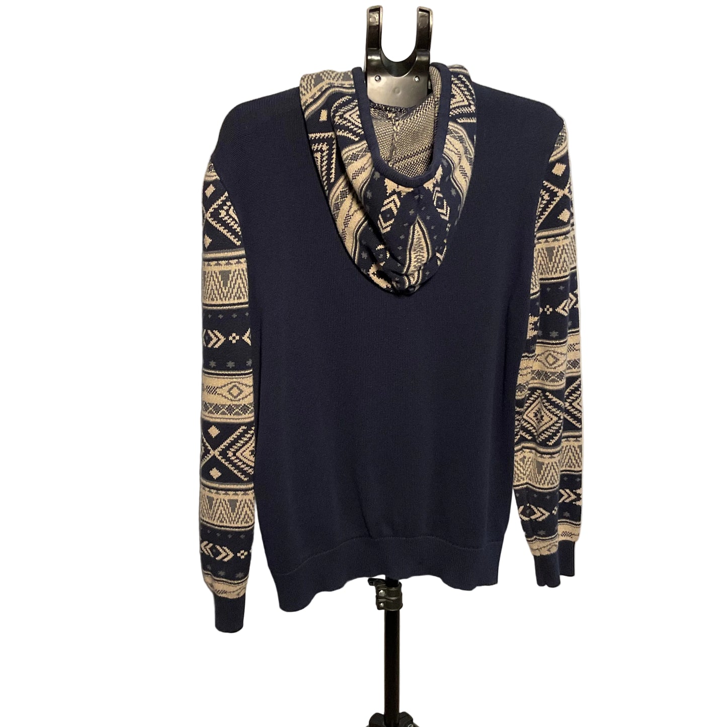 A navy blue crewneck sweater with beige and navy geometric Fair Isle patterns on the sleeves and a matching patterned cowl neck, displayed on a black hanger stand against a white circular backdrop.