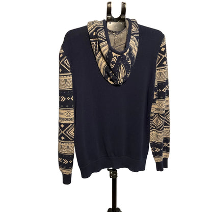 A navy blue crewneck sweater with beige and navy geometric Fair Isle patterns on the sleeves and a matching patterned cowl neck, displayed on a black hanger stand against a white circular backdrop.