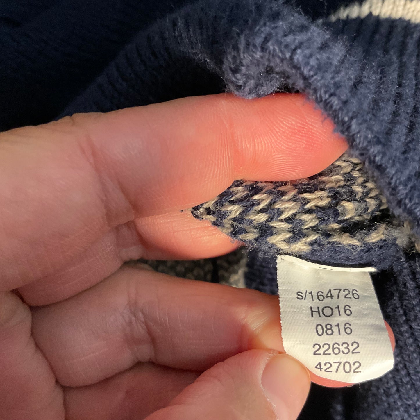 A close-up of a hand holding a small fabric tag sewn onto a navy blue and cream knit garment, showing printed size information.