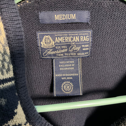 Close-up of a sweater's interior neck label reading 'MEDIUM' and 'American Rag,' with a dark navy knit collar and a mint green hanger visible.