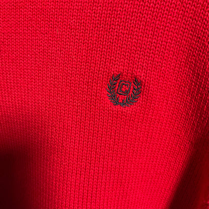 Close-up of a bright red knitted fabric with a small black embroidered logo in the center, showing a laurel wreath and square motif.