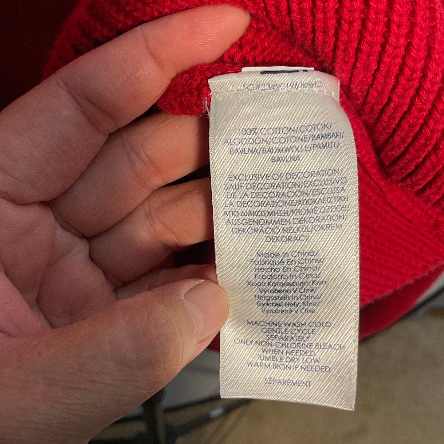 A close-up of a hand holding a white fabric care label attached to a bright red knit sweater.