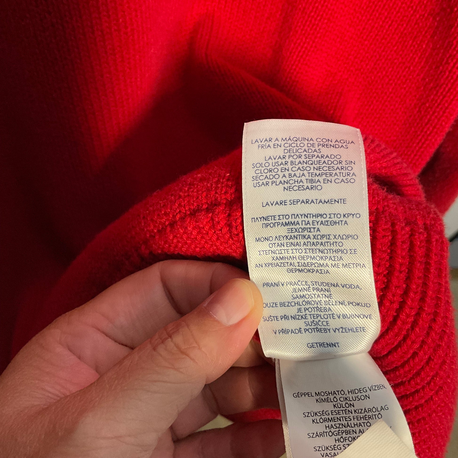 A close-up of a hand holding a white care label attached to a bright red knit garment, with the text printed in Spanish and Portuguese.