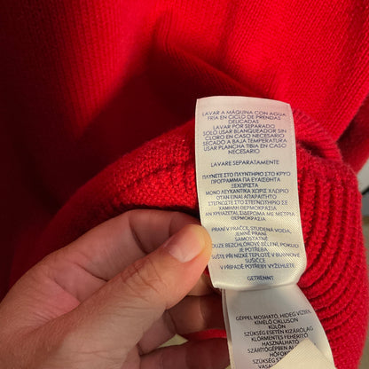 A close-up of a hand holding a white care label attached to a bright red knit garment, with the text printed in Spanish and Portuguese.