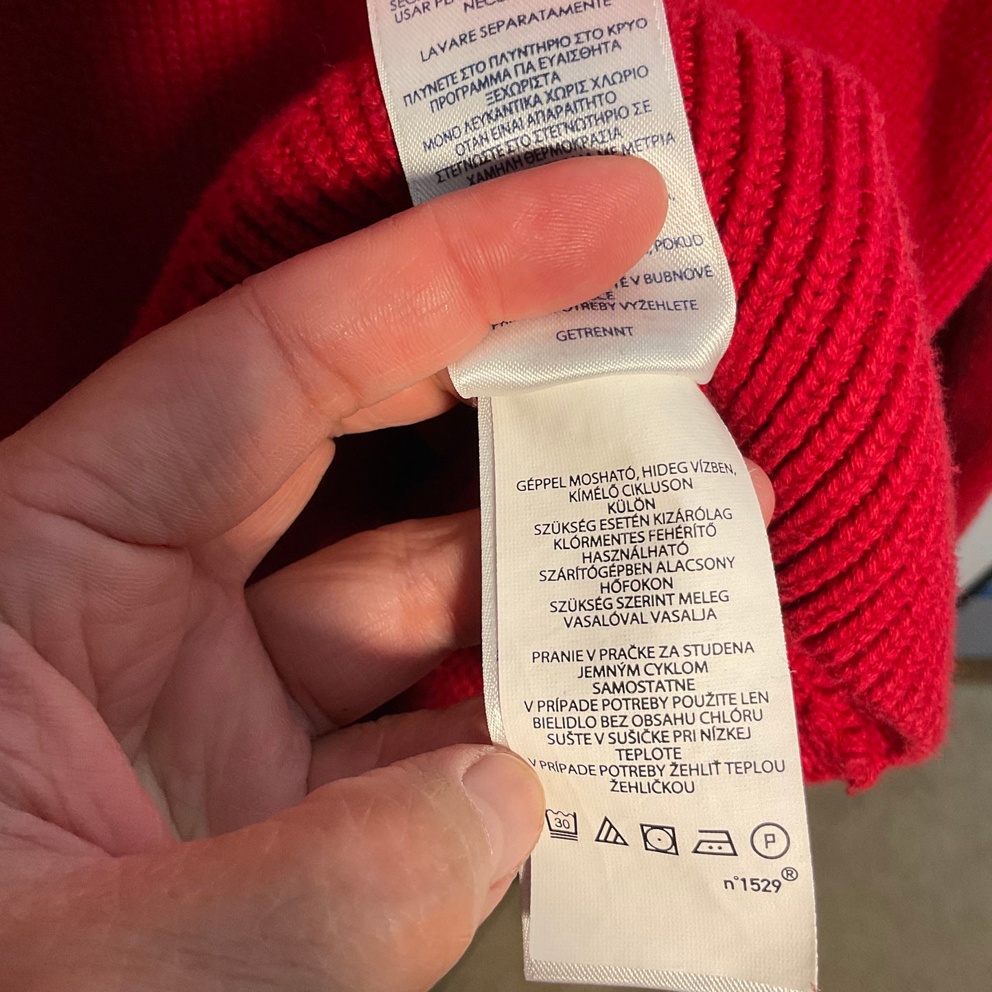 A close-up of a hand holding a white care instruction tag attached to a bright red knit garment, showing printed care instructions in multiple languages and washing symbols.