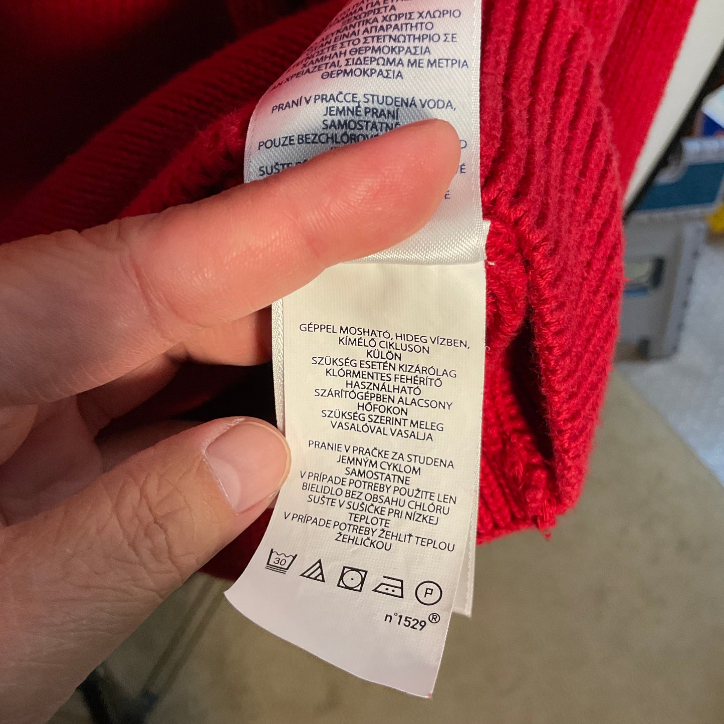 A close-up of a hand holding a white garment care label attached to a bright red knit sweater, with the label text partially visible in Czech.