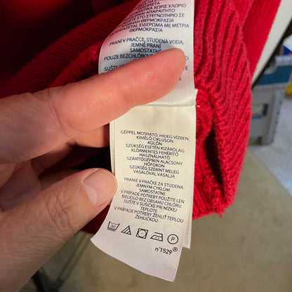 A close-up of a hand holding a white garment care label attached to a bright red knit sweater, with the label text partially visible in Czech.