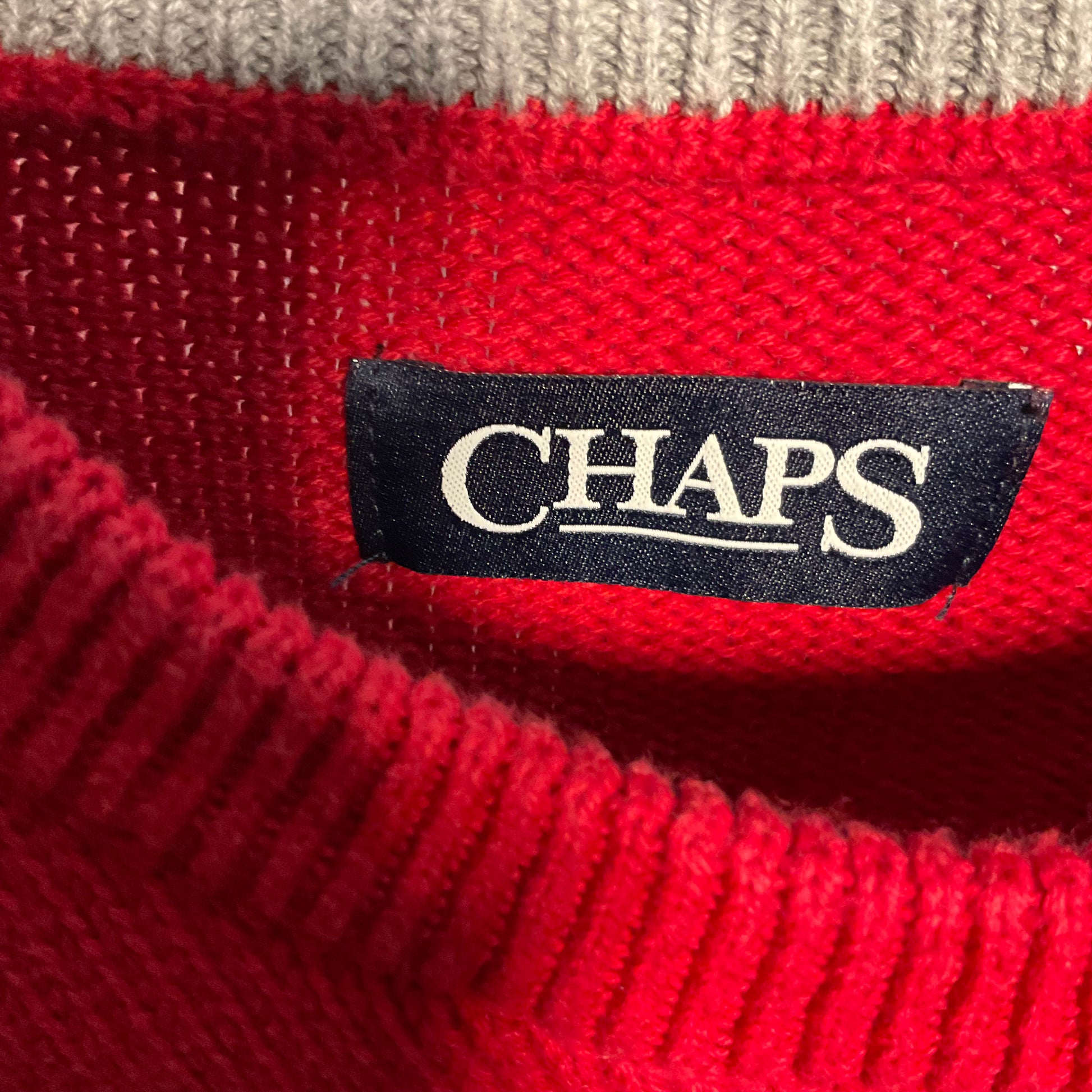 Close-up of a red knit sweater with a black CHAPS brand label sewn onto the inside collar, with a light beige ribbed knit band visible at the top.