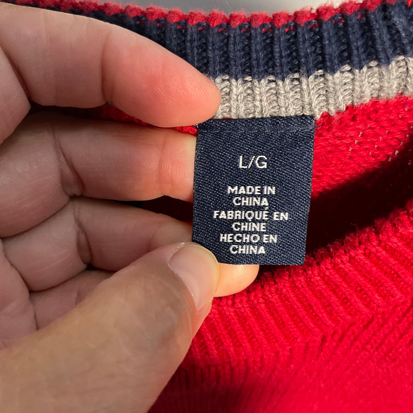 A close-up of a hand holding a navy blue size L/G tag on a red and navy striped knit sweater.