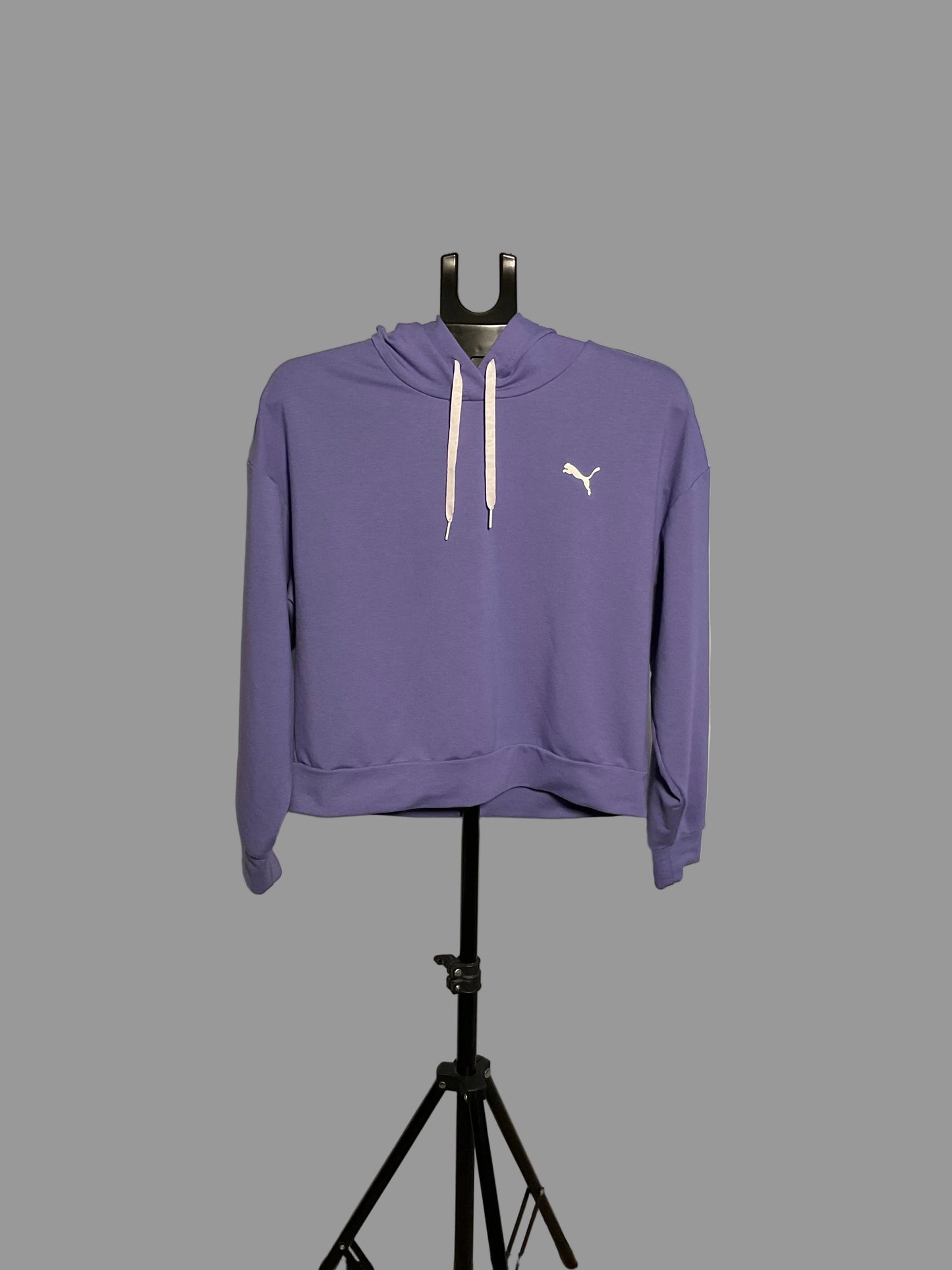 A purple Puma pullover hoodie with white drawstrings and a white cat logo on the chest, displayed on a black stand in front of a white circular studio backdrop.