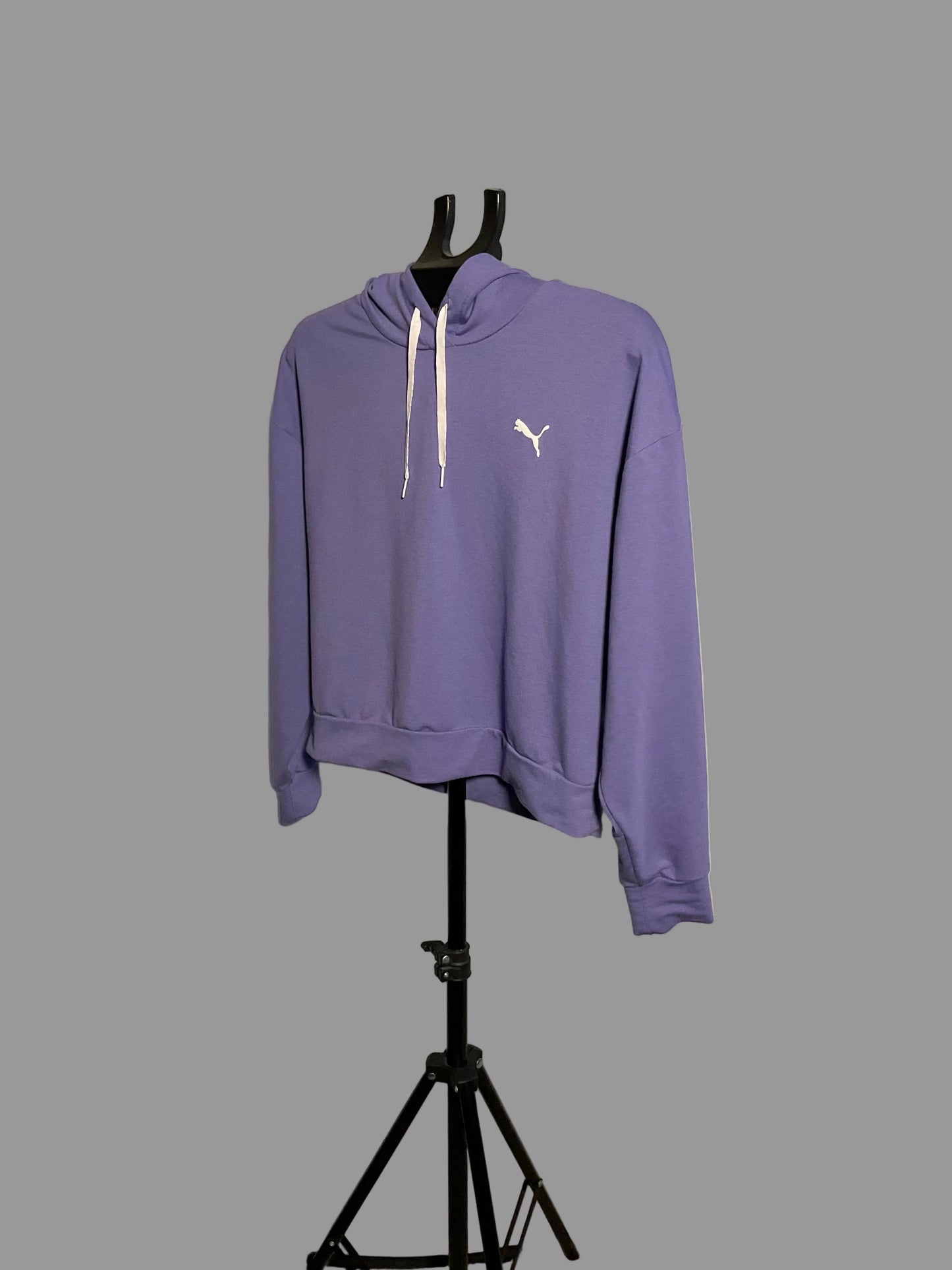 A purple Puma hoodie with white drawstrings and a white cat logo on the chest, displayed on a black hanger stand in front of a circular studio backdrop.