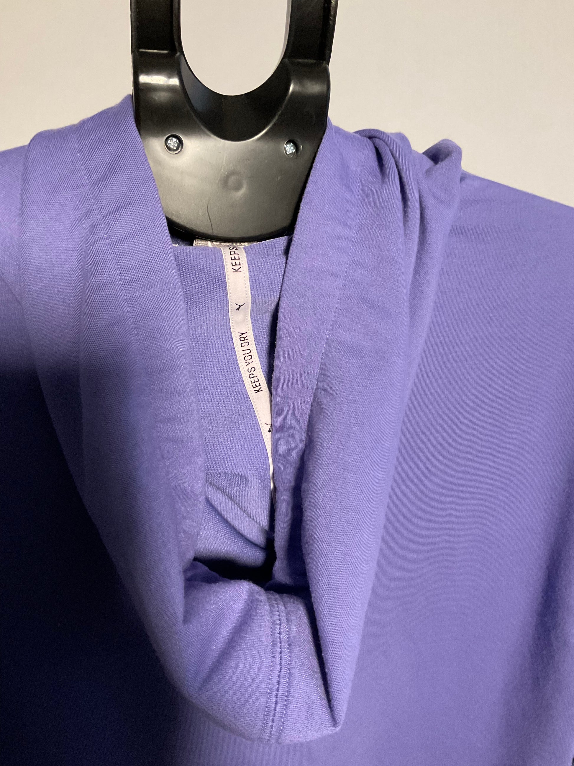 A close-up of a lavender-purple long-sleeve top draped on a black hanger, showing the soft texture of the fabric and a white inner neck label that reads 'AUGUST FAITH'.