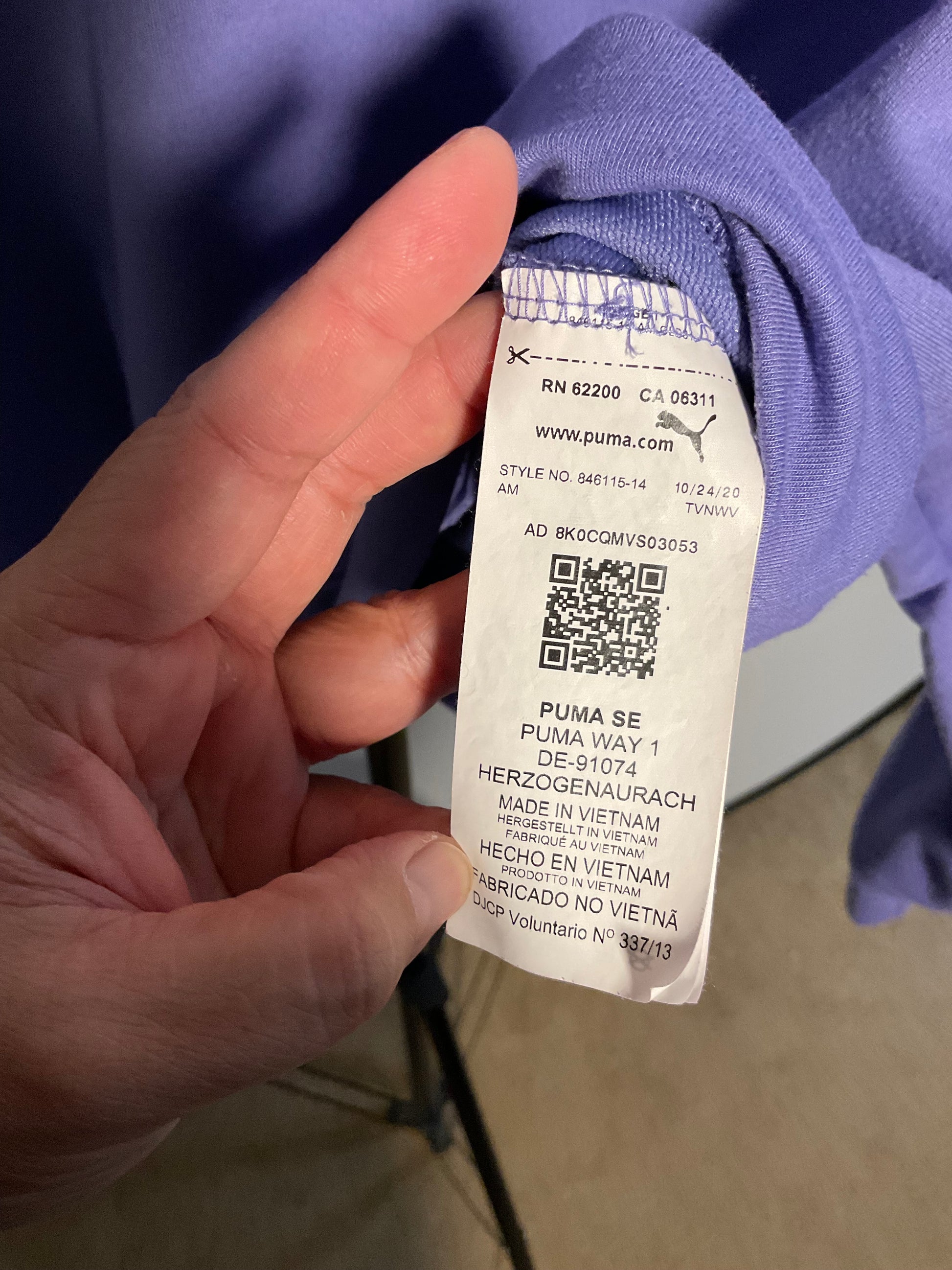 A close-up of a hand holding a white Puma garment tag attached to a purple sweatshirt, with the Puma brand name and website printed on the tag.