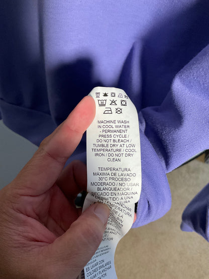 A close-up of a hand holding a white paper care label attached to a lavender-purple garment, with the garment filling most of the background.