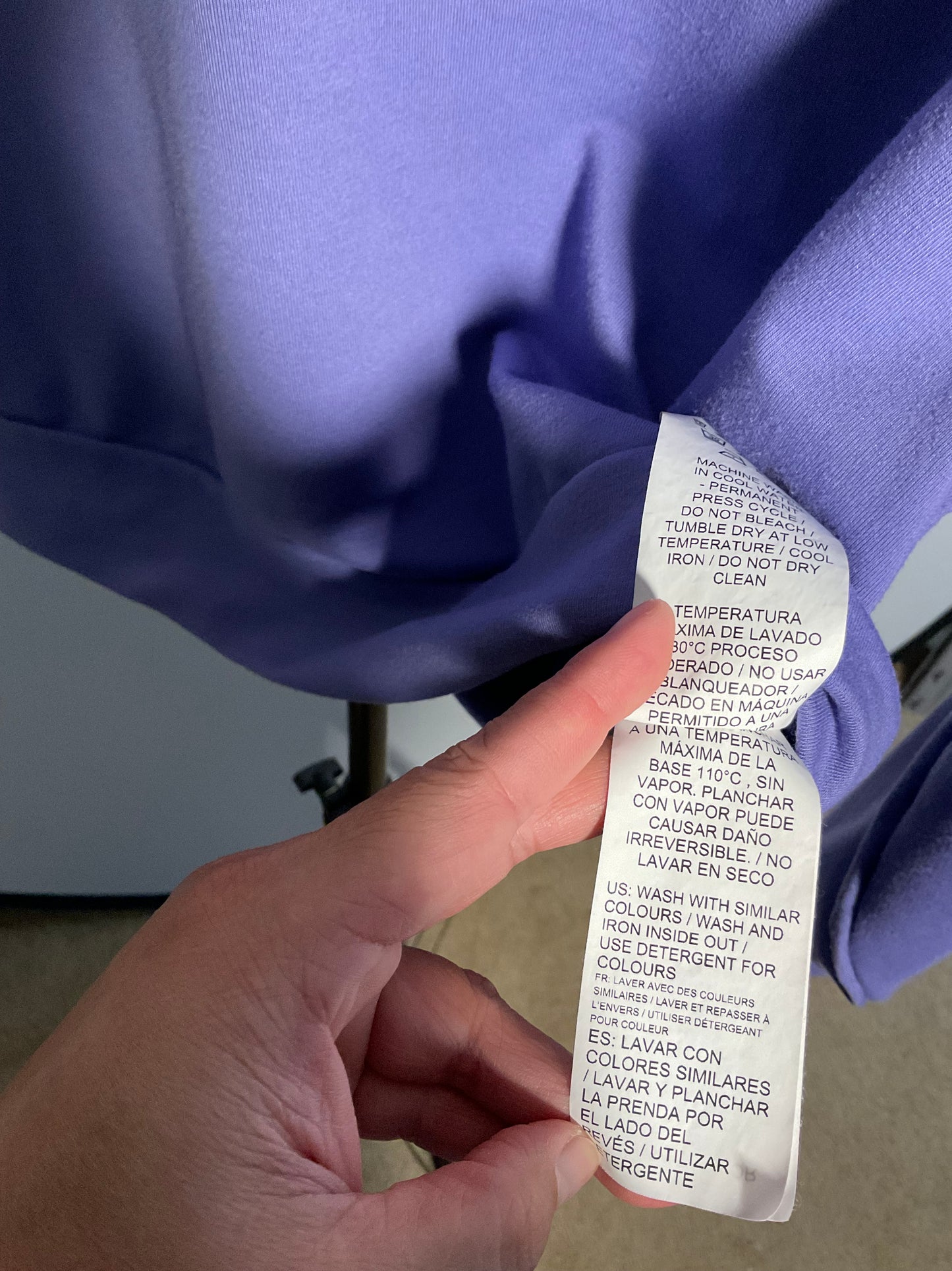 A close-up of a lavender-purple garment with a white care label being held by a hand. The label contains Spanish washing and drying instructions.