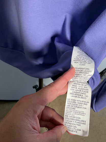 A close-up of a lavender-purple garment with a white care label being held by a hand. The label contains Spanish washing and drying instructions.
