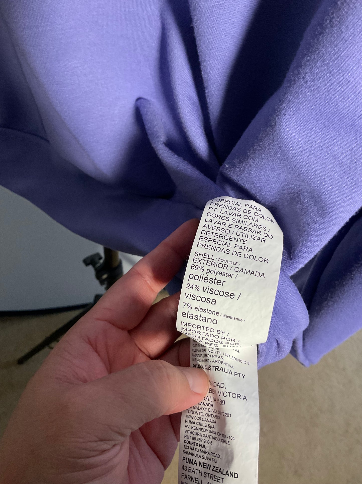 A close-up of a hand holding a white garment care label attached to a lavender-purple knit fabric garment.