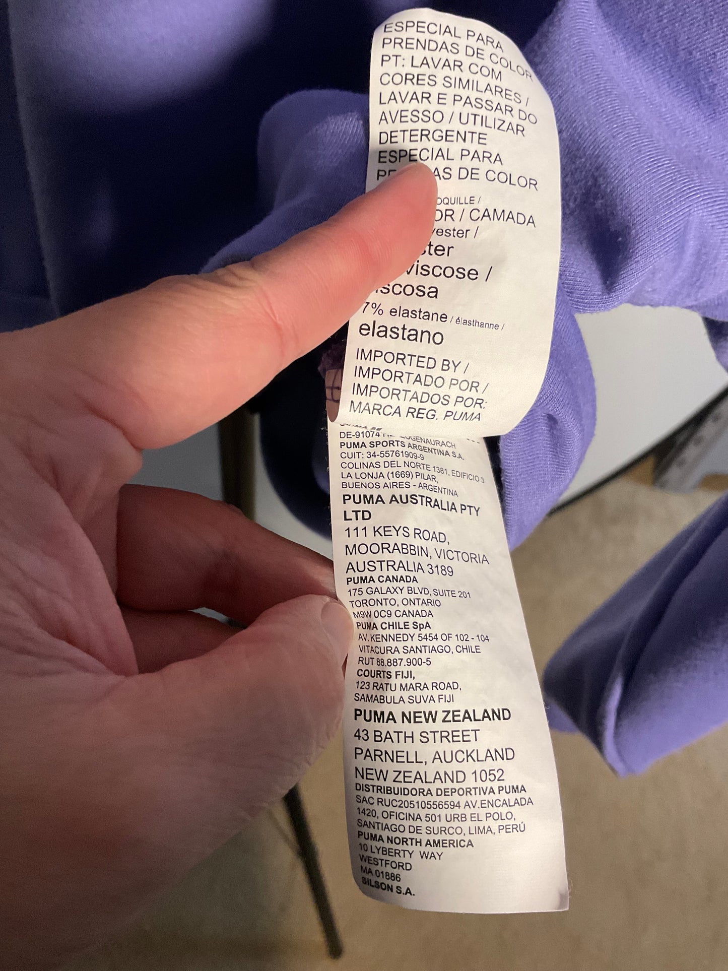 A close-up of a hand holding a white garment care label attached to a purple garment, showing printed text including Puma Australia Pty Ltd and care instructions.
