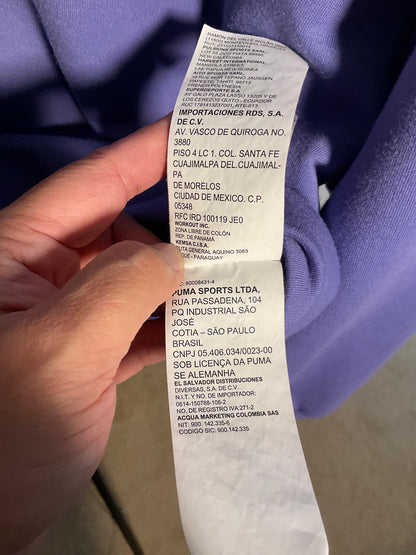 A close-up of a hand holding a white garment care label attached to a purple garment, showing printed company and shipping information in Spanish.