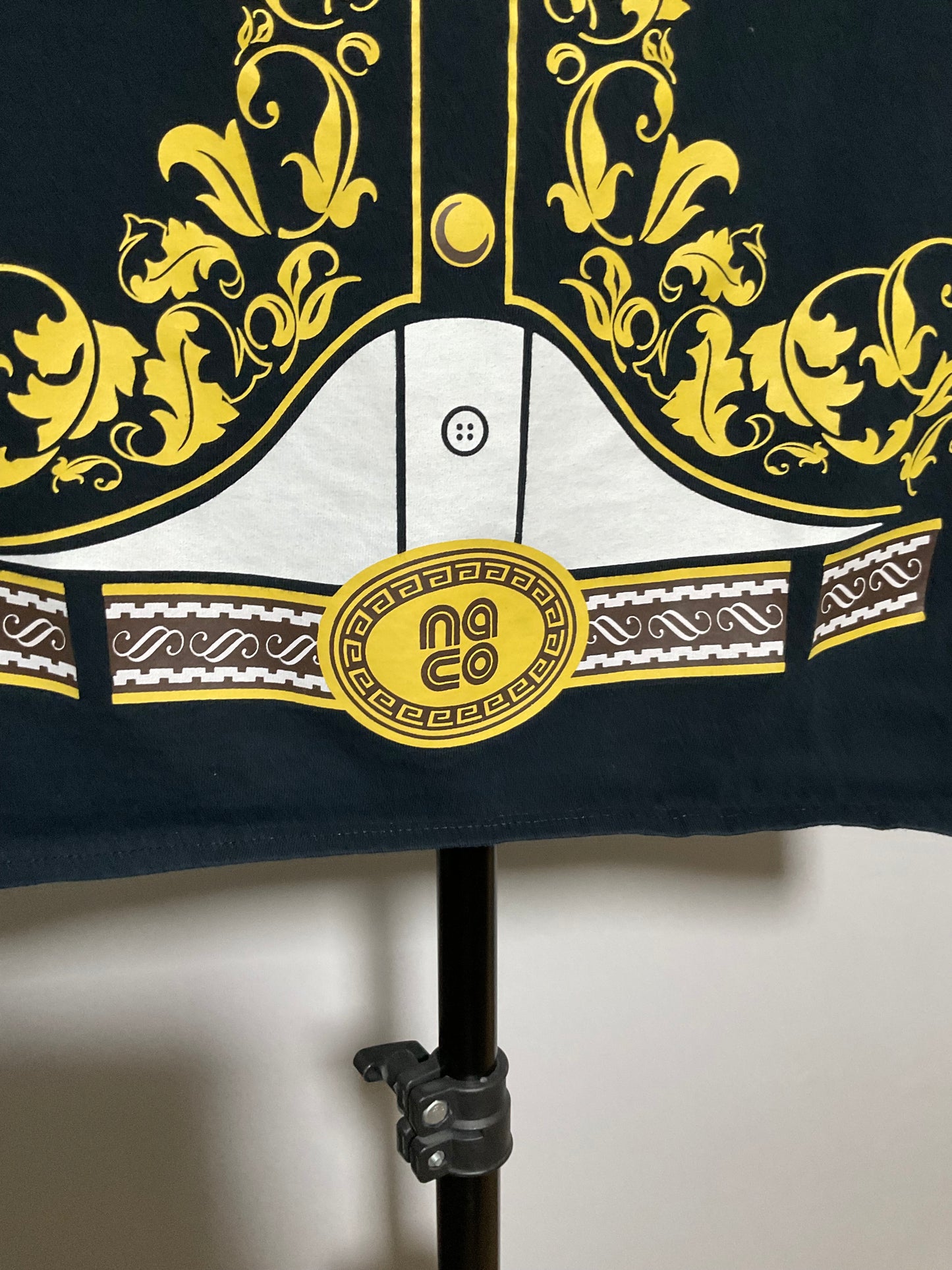 A close-up of a garment on a black stand, featuring a dark navy base with ornate yellow baroque-style scrollwork, a white central panel with a button motif, and a yellow circular patch with the text 'no co'.