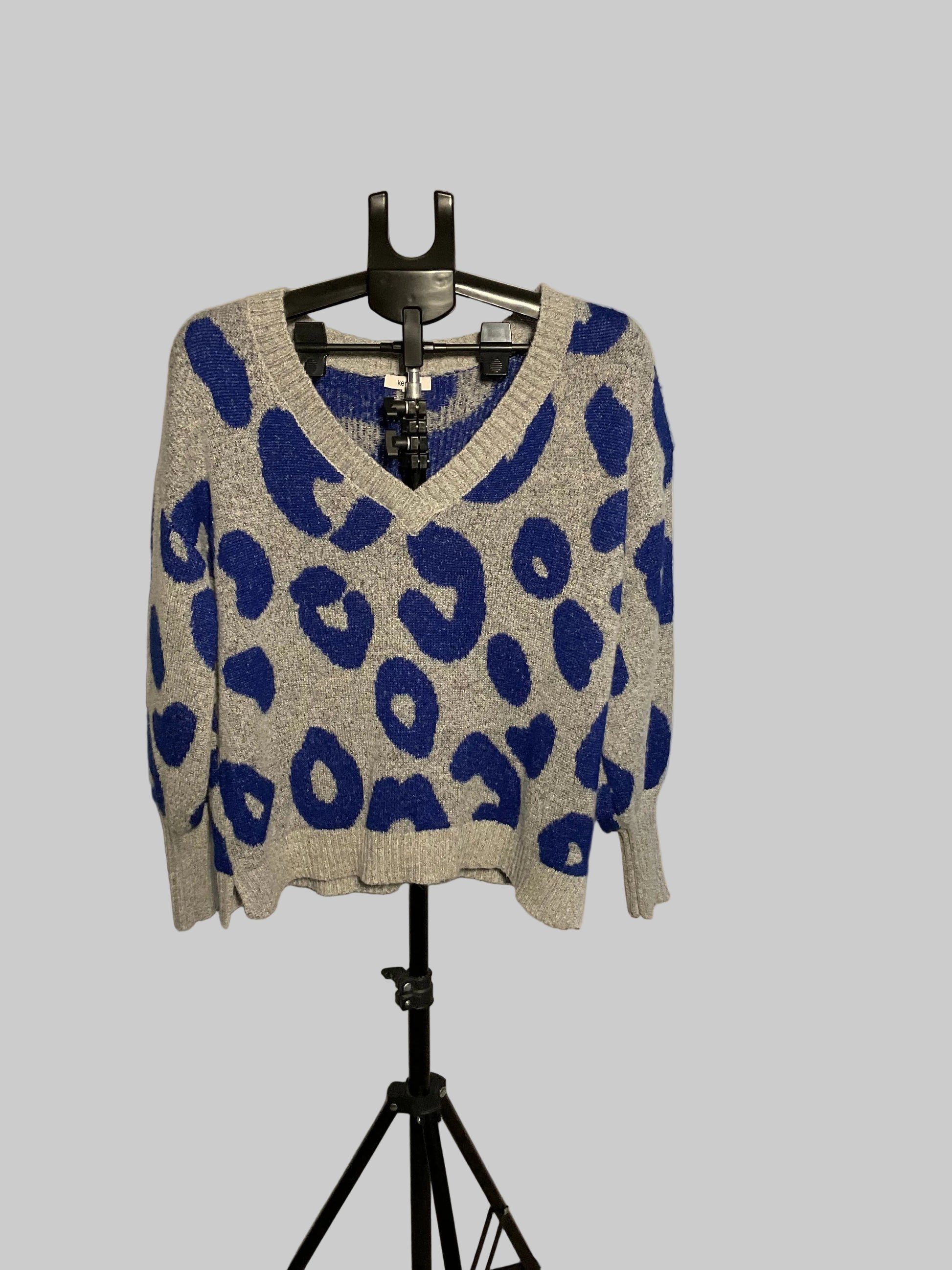 A gray knit sweater with a bold blue leopard print pattern, displayed on a black hanger stand in front of a large circular studio reflector.