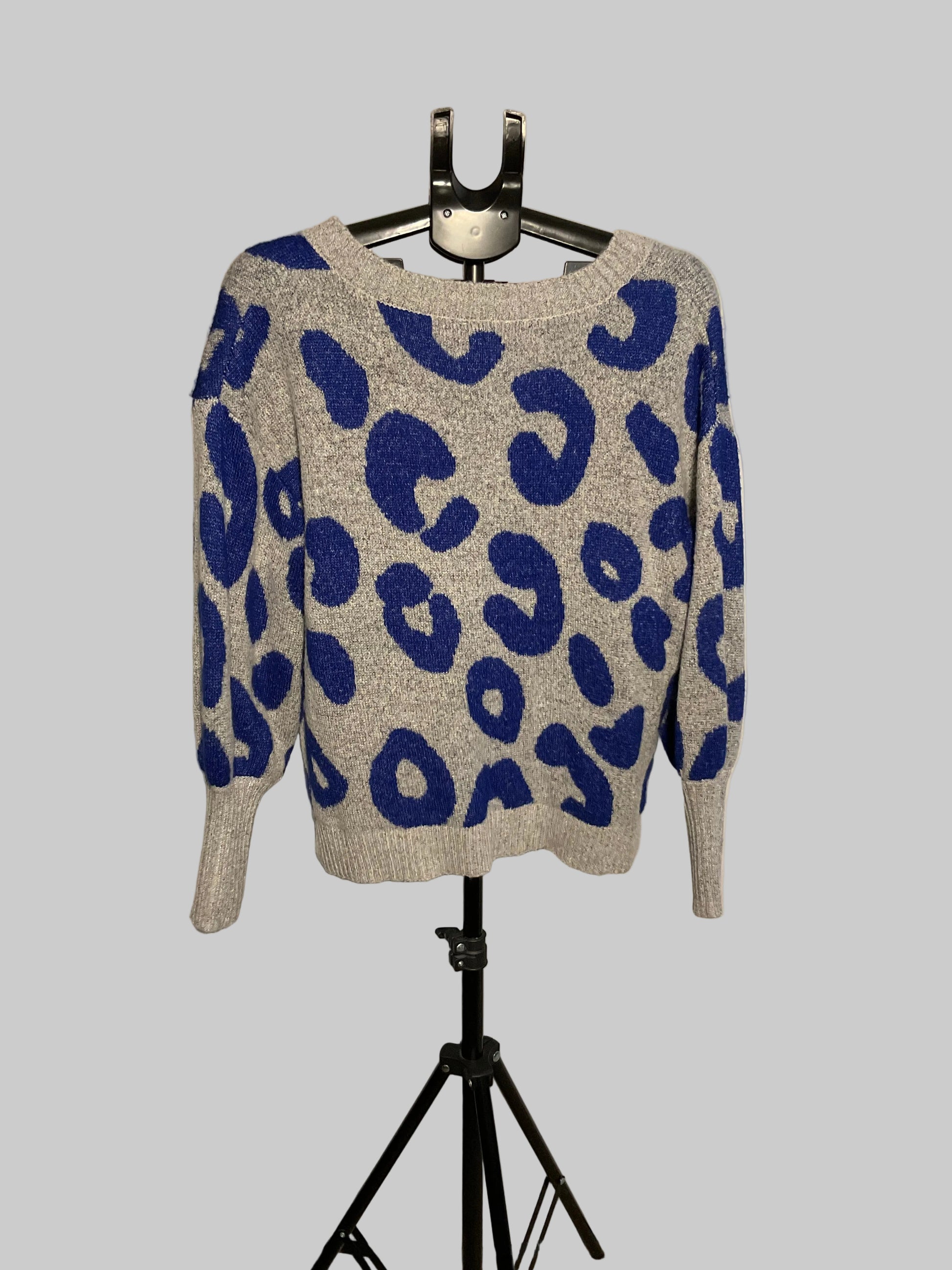 A gray knit sweater with a bold blue leopard print pattern, displayed on a black hanger stand against a white circular studio backdrop.