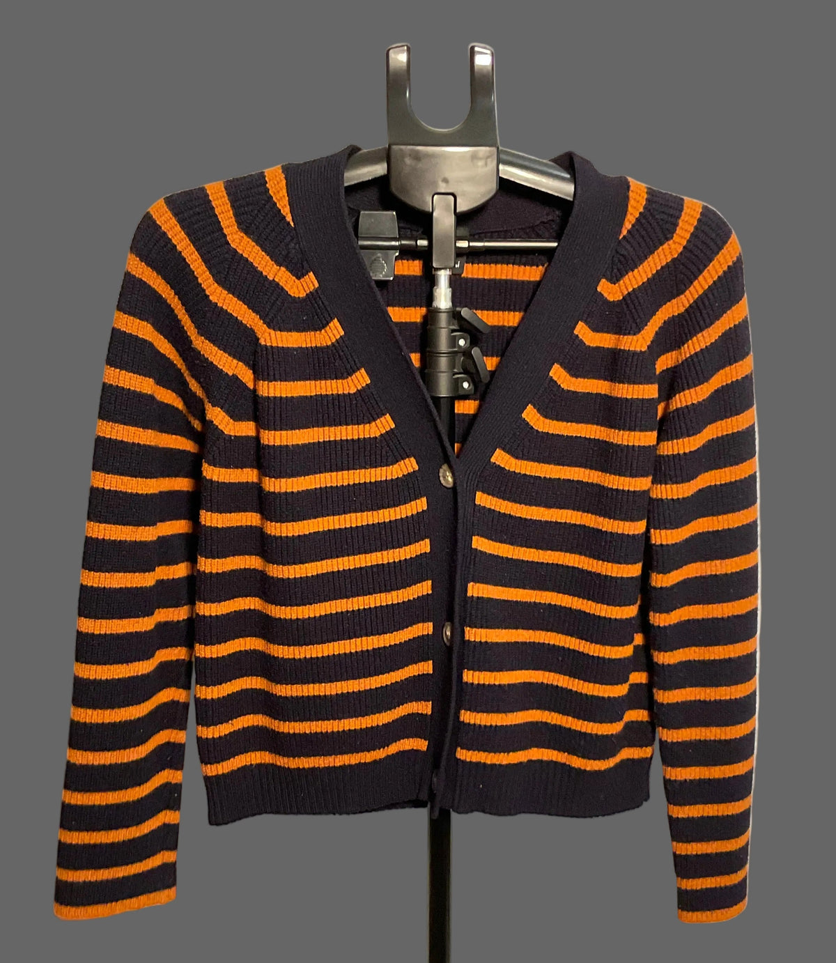 A cropped cardigan sweater with horizontal orange and black stripes, displayed on a black hanger stand against a white circular studio backdrop.