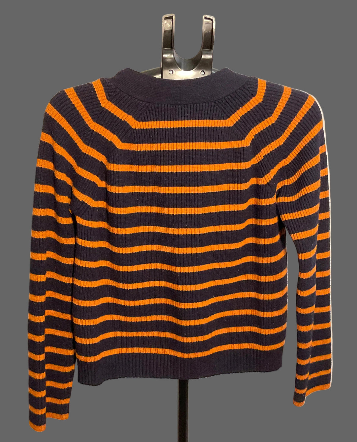 A long-sleeve knit sweater with horizontal navy blue and orange stripes, displayed on a black clothing hanger stand against a white circular backdrop.