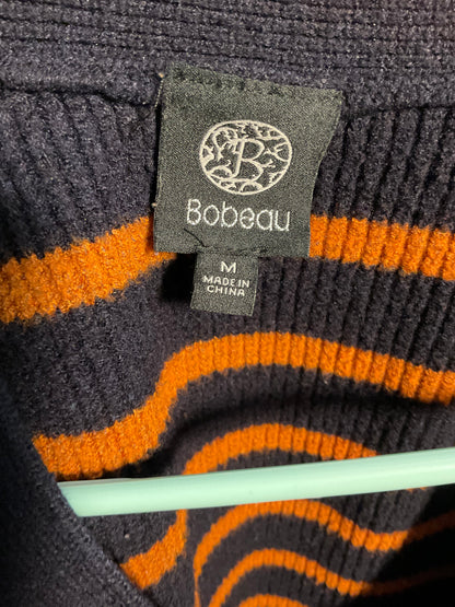 Close-up of a ribbed knit sweater label reading Bobeau, with a dark charcoal-gray fabric tag and a smaller size tag reading M / MADE IN CHINA. The sweater has wavy orange stripes on a dark charcoal-gray background.