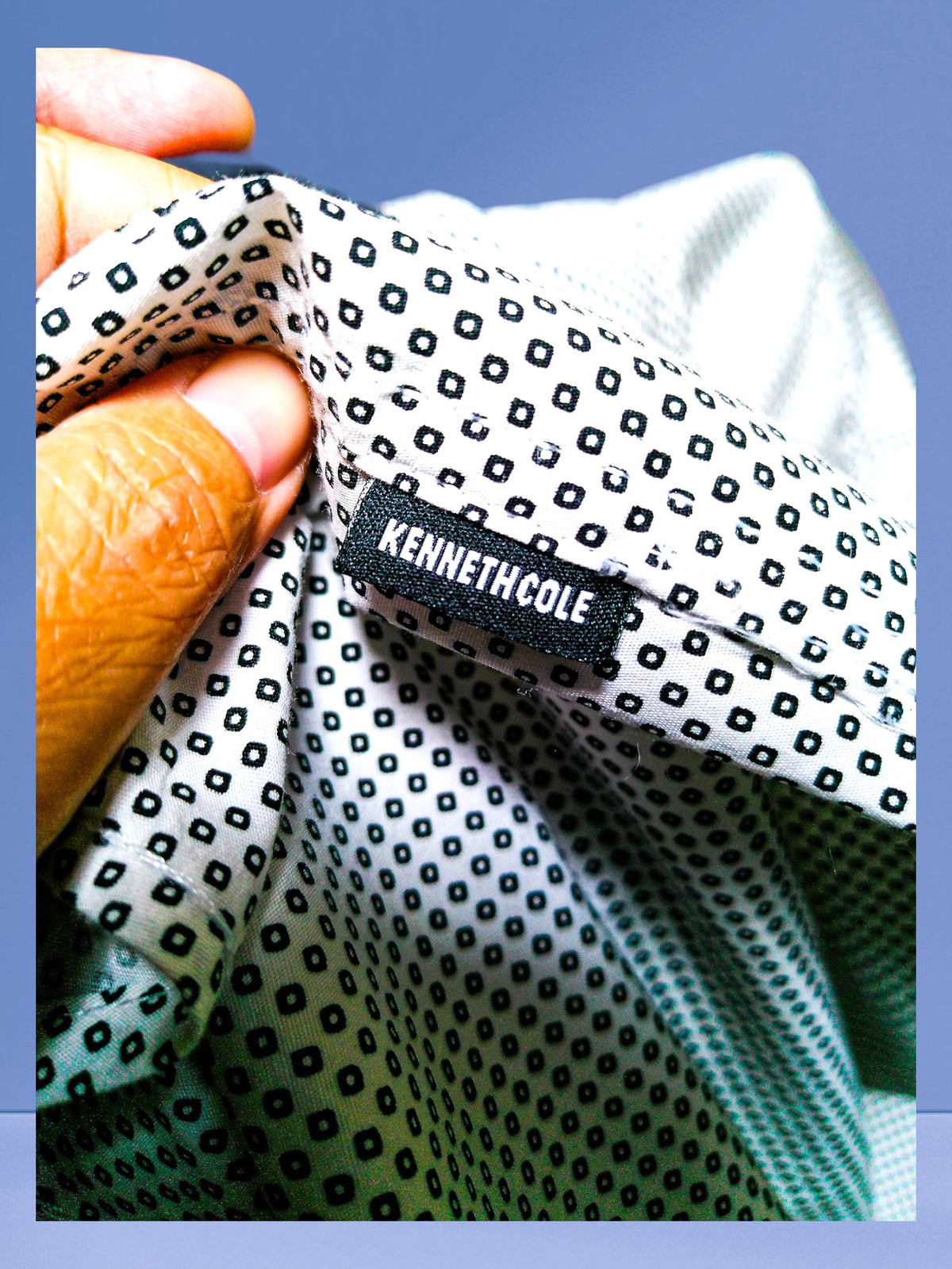 Close-up of a hand holding a folded piece of Kenneth Cole patterned fabric, showing the black brand label sewn onto the white and black micro-dot print.