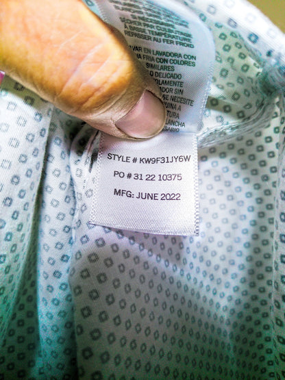 Close-up of a garment's interior neck label being held by a finger. The label reads 'STYLE # KW9F31JY6W PO # 31 22 10375 MFG: JUNE 2022' on a white fabric tag, with the garment interior showing a light teal and white geometric pattern.