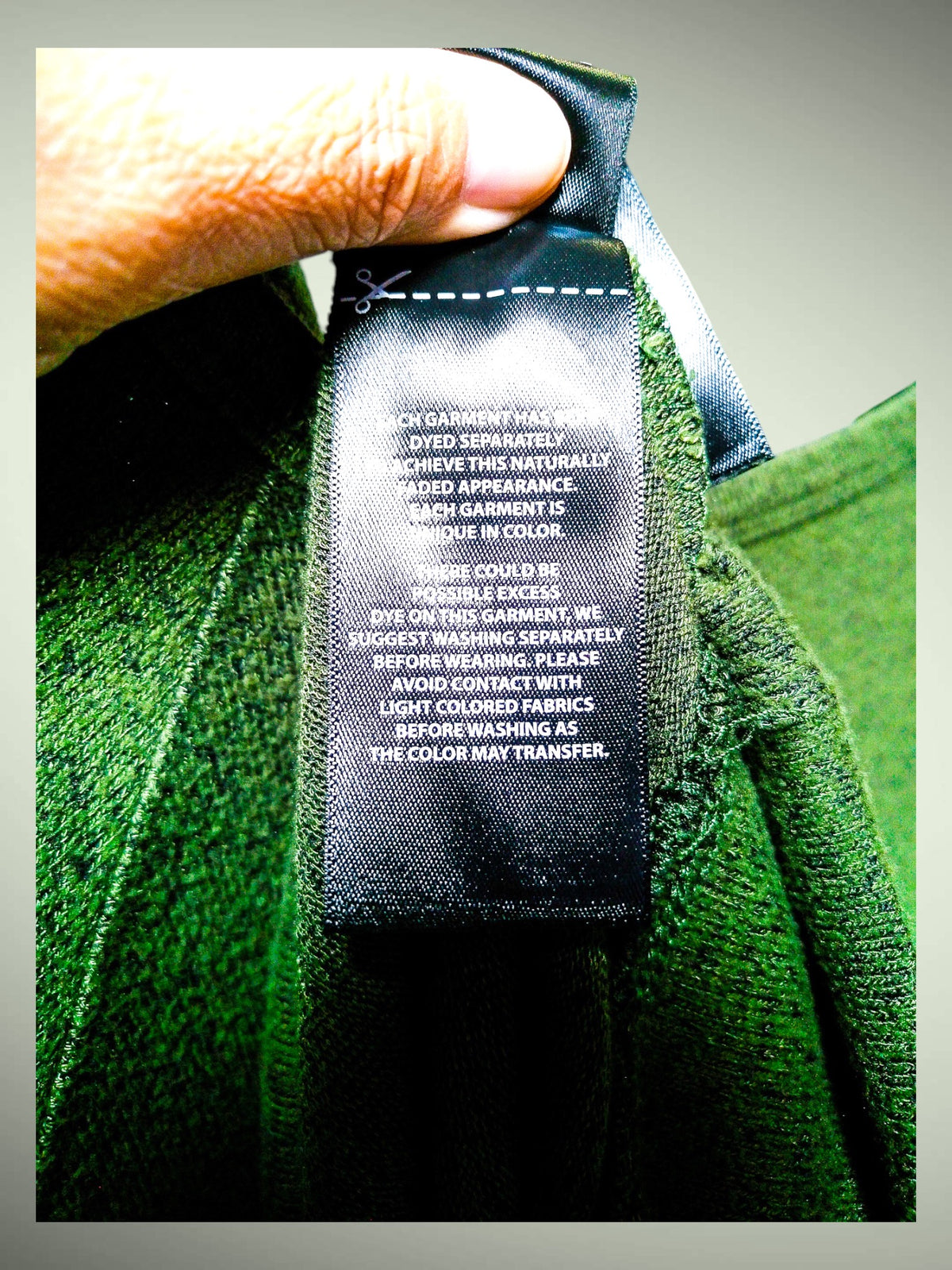 A close-up of a hand holding a black care label sewn onto the inside collar of a green garment. The label contains white text with care instructions, and the fabric around it is a rich green.